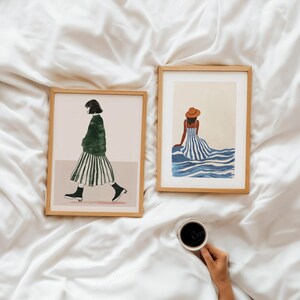 Puede incluir: Dos impresiones art&iacute;sticas enmarcadas sobre una superficie blanca. Una impresi&oacute;n representa a una persona con un su&eacute;ter verde y una falda a rayas caminando. La otra muestra a una persona con un vestido a rayas y un sombrero sentada cerca del agua azul. Una mano sostiene una taza de caf&eacute;.