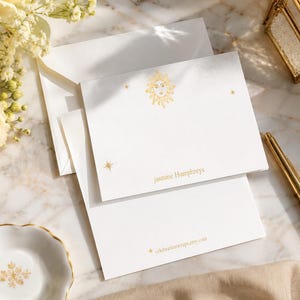 May include: White stationery set with gold accents. The set includes notecards, envelopes, and a pen. The notecards feature a sun illustration and the name "jasmine humphreys". The bottom card has the text "celebrationwrapsby.com".