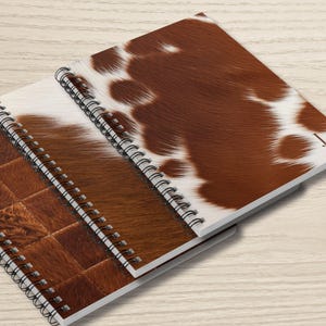 May include: Two spiral-bound notebooks with covers featuring a cowhide pattern. One notebook has a brown and white spotted design, while the other has a patchwork design in shades of brown. The notebooks are stacked on top of each other.