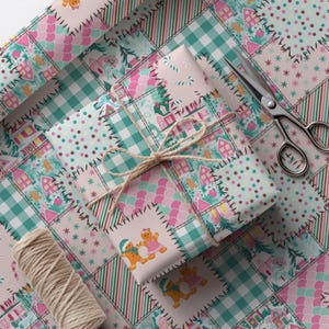Puede incluir: Un regalo envuelto con un diseño de patchwork con hombrecitos de jengibre, casas y copos de nieve. El papel de regalo es en tonos rosa, verde azulado y blanco, atado con hilo. También se ven unas tijeras y una bobina de hilo.