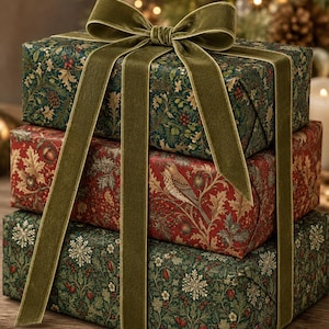 May include: A stack of three rectangular gift boxes wrapped in patterned paper featuring botanical motifs, berries, and a bird illustration. The boxes are tied together with a wide, olive green velvet ribbon tied in a large bow on top.