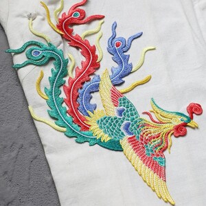 Puede incluir: Dise&ntilde;o de f&eacute;nix bordado en rojo, amarillo, azul y verde sobre tela blanca. El f&eacute;nix tiene alas y plumas de cola detalladas. El bordado es probablemente para decoraci&oacute;n de ropa o textiles.