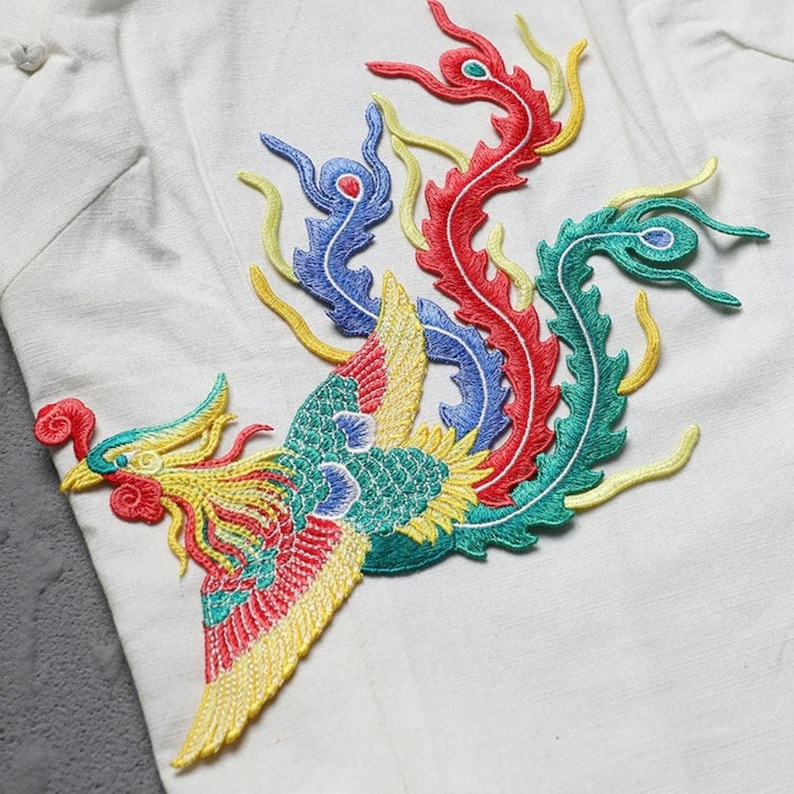 Puede incluir: Camisa blanca con un dise&ntilde;o de f&eacute;nix bordado colorido. El f&eacute;nix presenta hilos rojos, amarillos, azules y verdes, creando una imagen vibrante y detallada. La camisa tiene cuello mandar&iacute;n y cierre de botones.