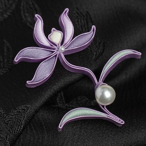 Könnte beinhalten: Lavendelfarbene Stoffblumenbrosche mit Perlenmitte und großem Perlenakzent. Die Brosche hat zwei grün umrandete Blätter vor einem schwarzen Hintergrund.