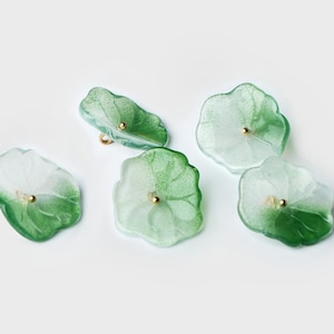 Puede incluir: Cinco botones verdes translúcidos con forma de pétalos de flor y bordes irregulares y ondulados. Cada botón presenta un efecto de degradado, pasando de un verde bosque intenso en la base a un blanco escarchado pálido en la parte superior, con un pequeño centro dorado.