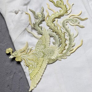 Puede incluir: Parche bordado con un dise&ntilde;o de f&eacute;nix dorado con detalles intrincados. El parche es en tonos dorados y verdes, con un ala y cola detalladas. El parche est&aacute; colocado sobre un fondo de tela blanca.