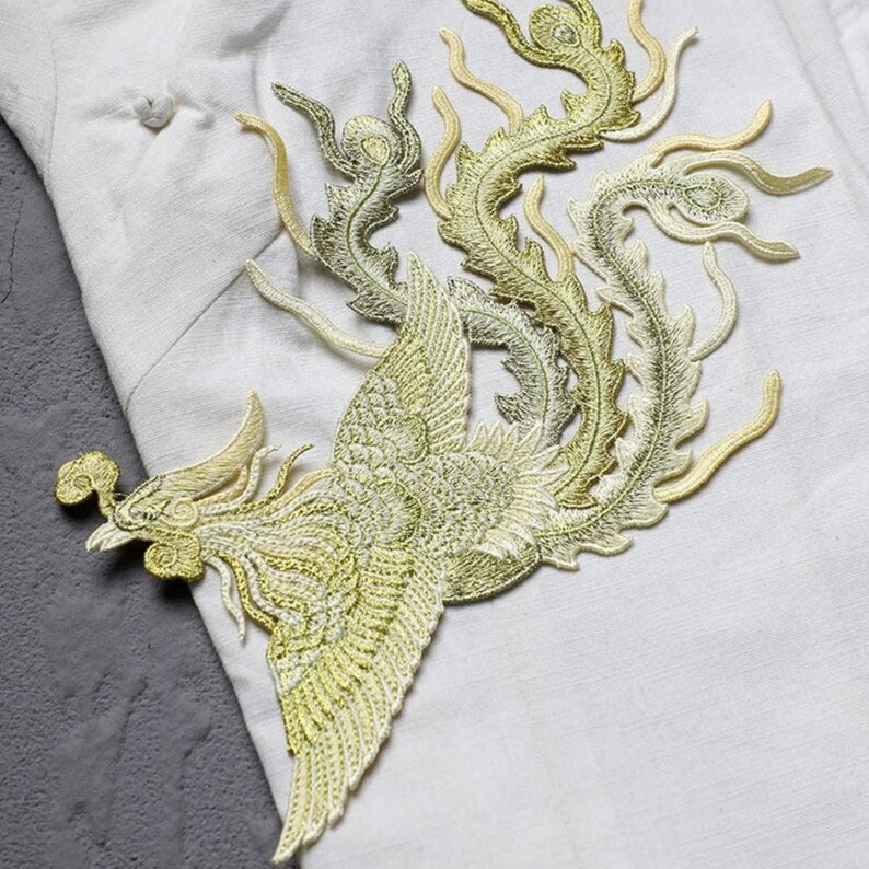 Puede incluir: Parche bordado con un dise&ntilde;o de f&eacute;nix dorado con detalles intrincados. El parche es en tonos dorados y verdes, con un ala y cola detalladas. El parche est&aacute; colocado sobre un fondo de tela blanca.