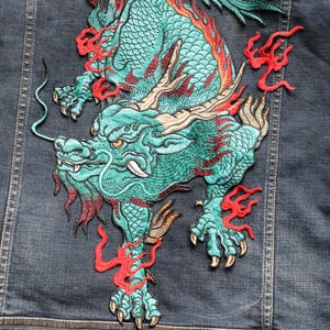 May include: A teal dragon embroidered patch with red flame details, set against a denim background. The dragon design features detailed stitching. This patch is suitable for customising clothing or accessories, adding a unique decorative element.