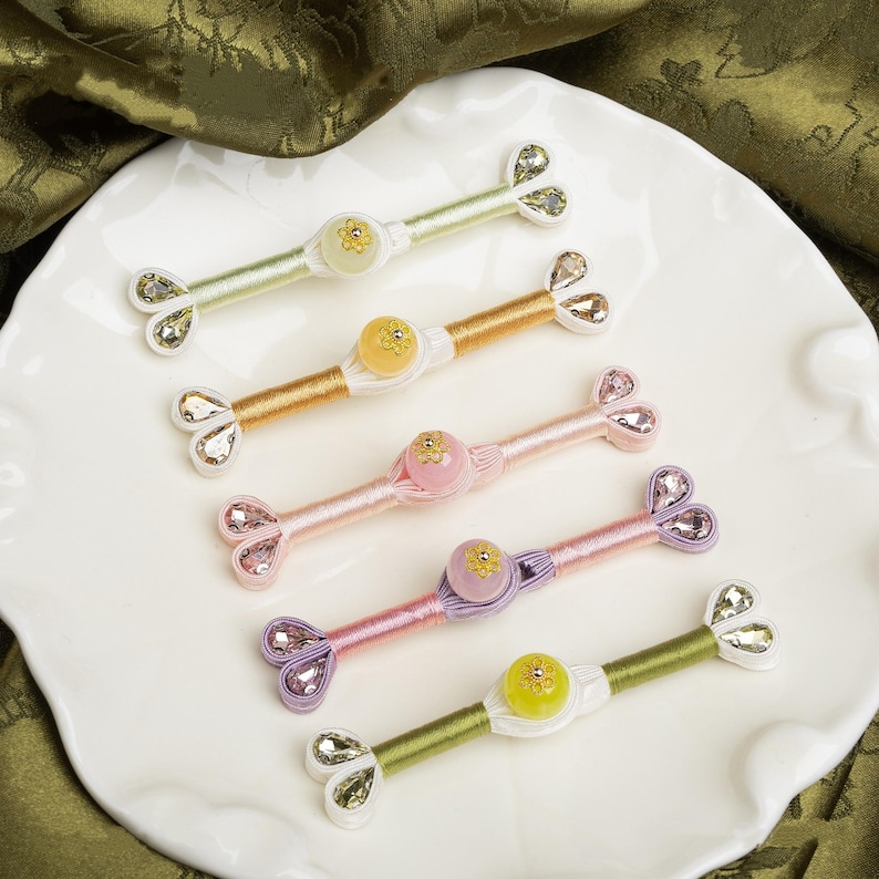 Op de afbeelding: Vijf kleurrijke, decoratieve haaraccessoires met een centrale kraal en bloemaccenten. Elk accessoire heeft een ander kleurenschema, waaronder roze, goud en groen, en is versierd met sprankelende versieringen. Ze zijn gerangschikt op een witte, geschulpte plaat.