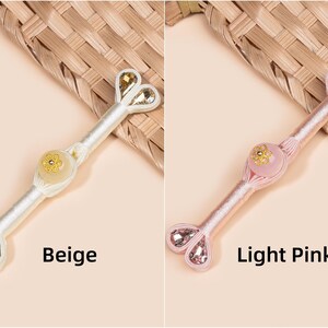 Op de afbeelding: Twee decoratieve haaraccessoires, &eacute;&eacute;n beige en &eacute;&eacute;n lichtroze, elk met een centrale bloemenversiering en hartvormige kristallen accenten. Het beige accessoire heeft goudkleurige kristallen, het roze heeft roze kristallen.