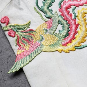 Puede incluir: Parche bordado de un f&eacute;nix en tonos amarillos, rosas, verdes y rojos. El parche est&aacute; cosido sobre una tela blanca, mostrando la detallada costura y los colores vibrantes del ave m&iacute;tica. El f&eacute;nix tiene una cabeza, alas y cola detalladas.