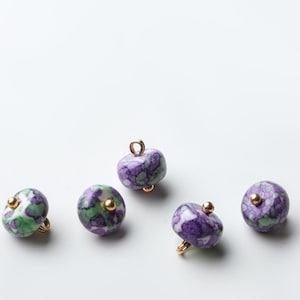 May include: Five gemstone beads with gold-colored findings. The beads are a mix of purple, green, and white, with a marbled appearance. Each bead has a small gold accent. The beads are round and appear to be for jewelry making.