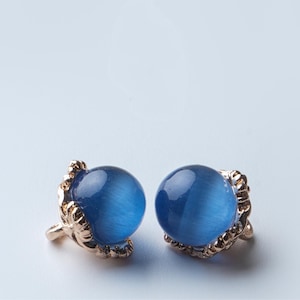 May include: A pair of stud earrings featuring a round, blue gemstone set in a gold-colored claw-like setting. The earrings have a polished, reflective surface and are displayed against a light blue background.