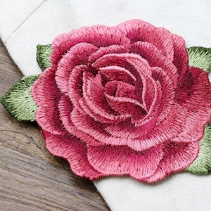 May include: Close-up of a detailed, embroidered rose in shades of pink and red, with green leaves. The floral design is set against a white fabric background, showcasing the intricate stitching and texture of the embroidery. The rose is a classic floral motif.