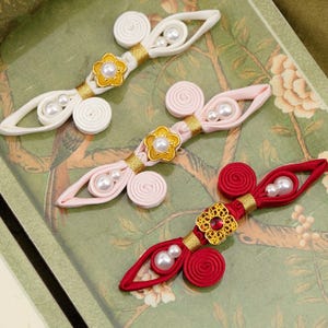 May include: Three decorative frog fasteners in white, light pink, and red. Each fastener features coiled fabric loops, gold-toned metallic accents, and central floral-shaped ornaments with embedded faux pearls or a red gemstone center.
