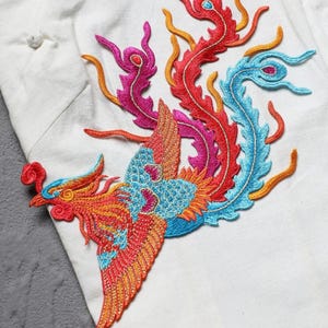 Puede incluir: Parche bordado de f&eacute;nix en colores vibrantes. El parche presenta un dise&ntilde;o detallado con hilos rojos, naranjas, azules y rosas. El f&eacute;nix se encuentra sobre un fondo de tela blanca, mostrando la intrincada costura y el arte del dise&ntilde;o.