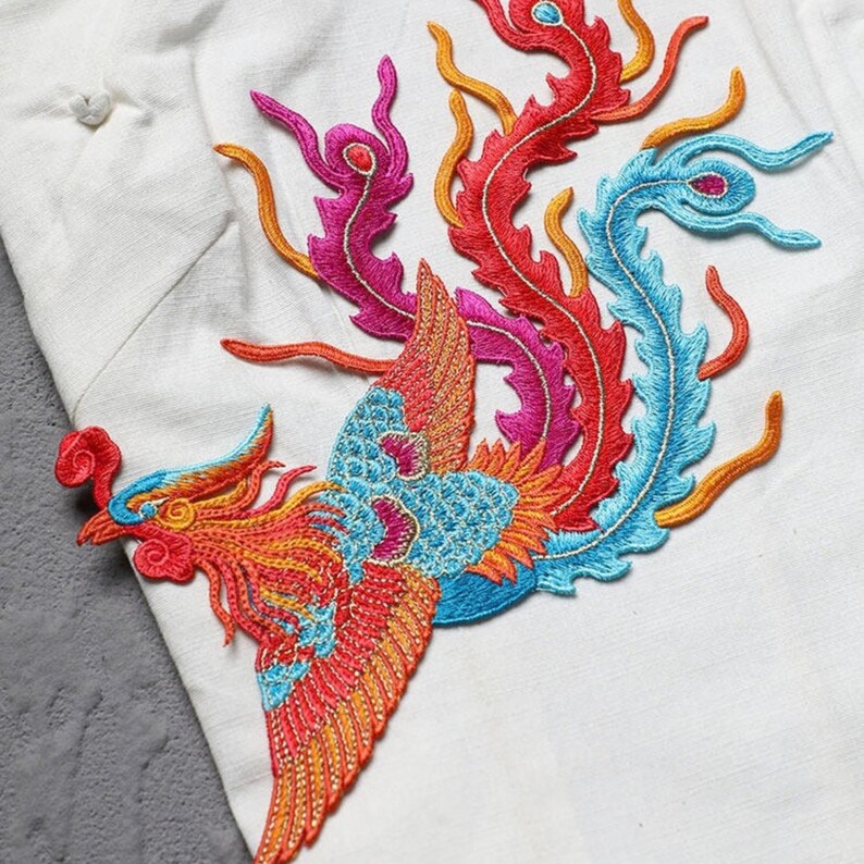 Puede incluir: Parche bordado de f&eacute;nix en colores vibrantes. El parche presenta un dise&ntilde;o detallado con hilos rojos, naranjas, azules y rosas. El f&eacute;nix se encuentra sobre un fondo de tela blanca, mostrando la intrincada costura y el arte del dise&ntilde;o.