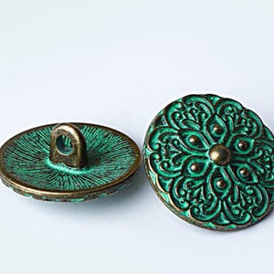 May include: Two antique-style buttons with a floral design. The buttons are teal and bronze, with a detailed pattern on the front and a loop on the back. The buttons are likely for sewing or crafts.