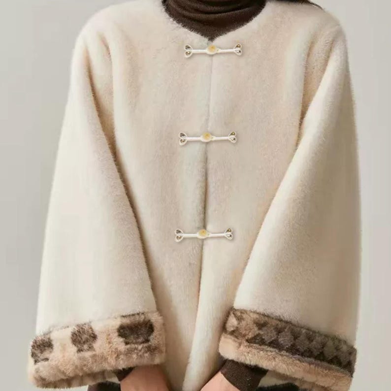 Op de afbeelding: Cr&egrave;mekleurige jas met lange mouwen en drie decoratieve sluitingen aan de voorkant. De jas heeft een zachte, getextureerde stof en contrasterende bruine en bruin/beige manchetten. De kraag is donkerbruin.