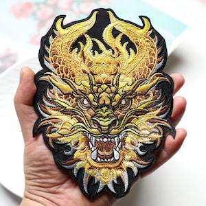 Puede incluir: Un parche bordado con un diseño detallado de cabeza de dragón. El dragón está representado en tonos dorados, amarillos y marrones con detalles blancos en los dientes y cuernos, sobre un fondo negro sólido con un borde negro.