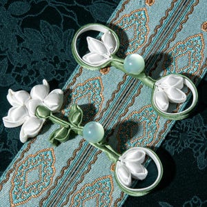 May include: Two decorative clasps with white flower and leaf accents, and a light green bead. The clasps are set against a teal and gold patterned fabric, with a dark teal background. The clasps are made of white satin ribbon and green satin ribbon.