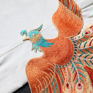 May include: An embroidered patch featuring a phoenix in flight. The bird has a bright orange body, a turquoise head and neck, and a long, detailed tail with gold, red, and turquoise accents. The embroidery shows intricate stitching throughout.