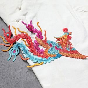 Puede incluir: Parche bordado de f&eacute;nix en colores vibrantes. El parche presenta un dise&ntilde;o detallado con hilos rojos, naranjas, azules y rosas. El f&eacute;nix se representa con las alas extendidas y una cola fluida, adecuado para ropa o proyectos de manualidades.