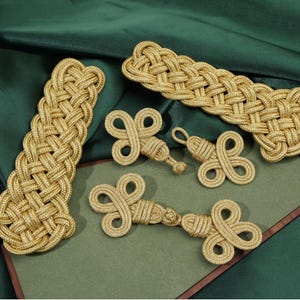 May include: A collection of gold-colored cord fasteners featuring intricate braided and knotted designs. The set includes one long rectangular braided piece, two smaller clover-shaped toggles, and one elongated toggle with two symmetrical knotted ends.