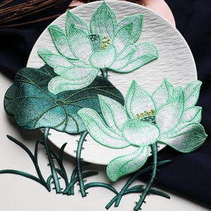 May include: An embroidered patch featuring two blooming lotus flowers and a large circular leaf. The design uses shades of white, mint green, and deep forest green thread. The lotus petals have gradient coloring, and the stems are thin and dark green.