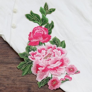 May include: Embroidered floral applique featuring pink and fuchsia peony blooms with green leaves. The applique is on a white fabric background, with a wooden surface visible in the lower portion of the image. The design includes three small pink flowers.