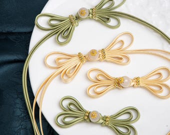 Op de afbeelding: Vier decoratieve sluitingen van satijnkoord in strikvorm. Elke sluiting heeft een centrale doorschijnende kraal met een klein goudkleurig bloemaccent en gouden metalen kralen aan weerszijden. Twee zijn saliegroen en twee zijn crèmekleurig.