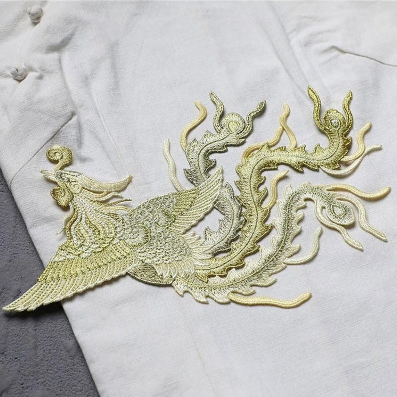 Puede incluir: Parche bordado con un dise&ntilde;o de f&eacute;nix en tonos dorados y verdes. El parche est&aacute; cosido sobre tela blanca, resaltando los detalles de las plumas y la cola fluida del ave. El dise&ntilde;o es intrincado.
