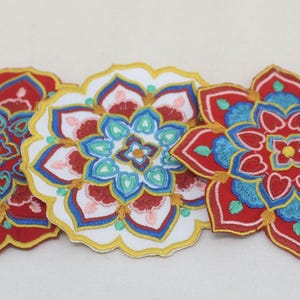 Puede incluir: Primer plano de tres parches florales bordados. Los parches presentan un diseño simétrico con colores rojo, blanco, azul y dorado. Los parches son probablemente para fines decorativos, como adornar ropa o accesorios.