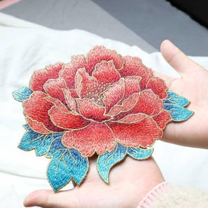 May include: A large, embroidered floral applique featuring a peony design. The flower has layers of red petals with gold thread outlining and a gold center. Blue leaves with gold trim accent the flower. The applique is held in a person's hands.