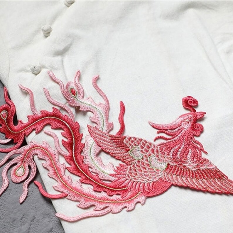 Puede incluir: Parche bordado con un f&eacute;nix estilizado en tonos rojos y rosas. El dise&ntilde;o presenta detalles intrincados y acentos de hilo dorado, sobre un fondo de tela blanca. El f&eacute;nix se representa con las alas extendidas y plumas de cola fluidas.