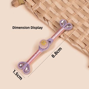 Op de afbeelding: Decoratieve roze en lavendelkleurige knoop met een bloemmotief en heldere kristallen accenten. De knoop is 8,8 cm lang en 1,5 cm breed. De tekst "Dimension Display" is zichtbaar.