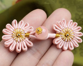 Op de afbeelding: Close-up van twee roze bloemvormige stoffen knopen met gouden accenten. Elke bloem heeft een centraal gouden ontwerp en is verbonden door een roze koord met een centrale roze kraal. De knopen worden in een hand gehouden.