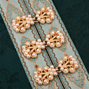 Può includere: Tre chiusure color oro con accenti di perle finte sono esposte su un tessuto fantasia turchese e verde. Le chiusure hanno un design floreale ornato. Lo sfondo è un tessuto verde scuro.
