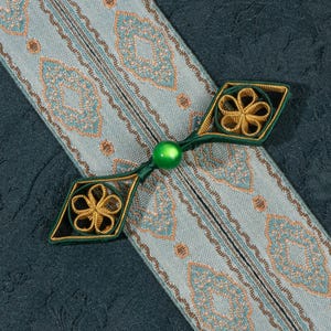 Könnte beinhalten: Nahaufnahme eines dekorativen Knopfes und Stoffes. Der Knopf ist grün und goldfarben, mit einem floralen Design. Der Stoff ist hellblau mit einem sich wiederholenden Muster in Gold und Türkis. Der Hintergrund ist ein dunklerer teal-farbener Stoff.