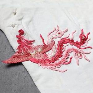 Puede incluir: Parche bordado con dise&ntilde;o de f&eacute;nix en tonos rojos y rosas. El ave, con alas detalladas y cola fluida, destaca sobre un fondo de tela blanca. Ideal para decorar prendas o accesorios.