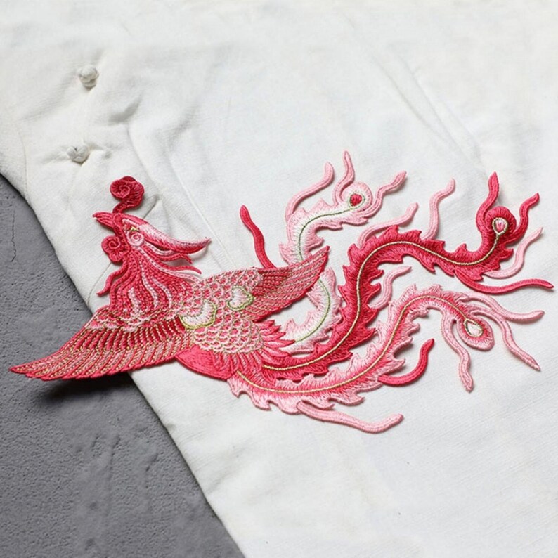 Puede incluir: Parche bordado con dise&ntilde;o de f&eacute;nix en tonos rojos y rosas. El ave, con alas detalladas y cola fluida, destaca sobre un fondo de tela blanca. Ideal para decorar prendas o accesorios.