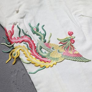 Puede incluir: Parche bordado con un dise&ntilde;o de f&eacute;nix colorido. El parche muestra un p&aacute;jaro detallado con hilos rojos, rosas, amarillos y verdes. El parche est&aacute; cosido sobre una tela blanca, con la cola del dise&ntilde;o curv&aacute;ndose sobre el material.