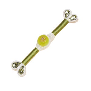 Op de afbeelding: Een decoratief accessoire met een centrale lichtgroene kraal versierd met een goudkleurig bloemmotief. Het stuk heeft olijfgroene en witte accenten, met sprankelende, heldere, hartvormige versieringen aan elk uiteinde. Het ontwerp is symmetrisch.