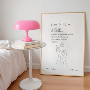 K&ouml;nnte beinhalten: Eine pinkfarbene Pilzlampe steht auf einem wei&szlig;en Beistelltisch mit B&uuml;chern. Ein gerahmter Druck mit dem Text "ONCE UPON A TIME..." und "throne of glass" lehnt an der Wand. Der Raum hat einen minimalistischen Stil.