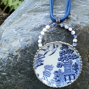 May include: A necklace featuring a circular pendant made from a piece of blue and white willow pattern ceramic china. The pendant is framed in silver metal and topped with a semi-circle of blue and white stone beads on a blue cord necklace.