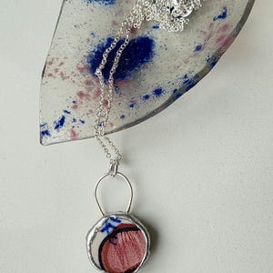 Peut inclure: Un collier en argent avec un pendentif circulaire. Le pendentif présente un motif abstrait rouge et blanc, encadré par une lunette en argent. Le collier repose sur un plat décoratif en verre avec des éclaboussures bleues et roses.