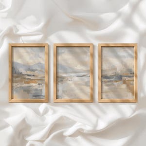 Puede incluir: Un conjunto de tres piezas de arte mural rectangulares verticales en marcos de madera clara. Cada marco contiene una pintura de paisaje abstracta con tonos apagados de beige, crema, gris suave y marrón tierra con texturas de pinceladas visibles.