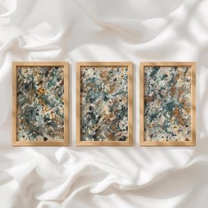 Può includere: Un set di tre quadri rettangolari con cornici in legno chiaro. Ogni cornice contiene un dipinto astratto caratterizzato da un caotico motivo a schizzi nei colori verde acqua, verde scuro, giallo senape, bianco e beige su fondo neutro.