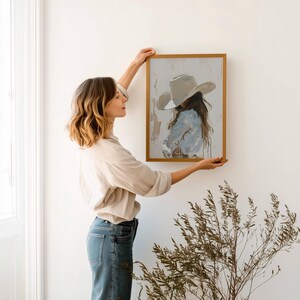 Pode incluir: Uma obra de arte emoldurada com uma pintura de uma pessoa usando um chap&eacute;u de cowboy e jaqueta jeans. A obra de arte tem uma paleta de cores claras e uma moldura dourada. A pessoa est&aacute; segurando a obra contra uma parede branca.