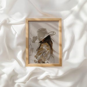 May include: A framed art print featuring a stylised illustration of a figure wearing a wide-brimmed hat. The artwork uses a neutral colour palette of beige, cream, and brown tones to depict the subject from the back, with long hair flowing.